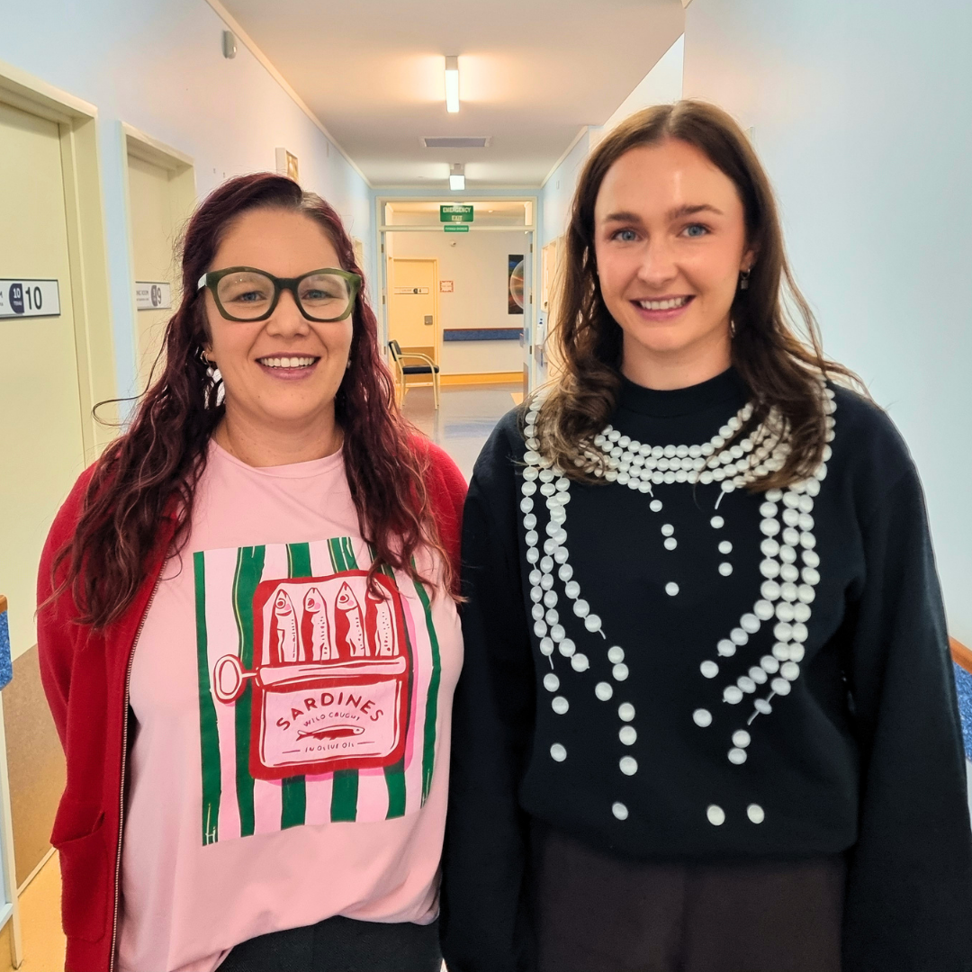 Two smiling women (Lauren and Morgan, Carefirst pharmacist prescribers) stand in a bright hallway; one wears glasses, a red cardigan and sardine-print shirt, the other a black sweater with a bead print on the front.